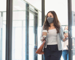 A mid adult businesswoman enters her office during the COVID-19 pandemic. A colleague is walking behind her, practicing the proper social distancing protocol. The businesswomen are wearing protective face masks.