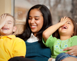 A nanny smiles warmly as a young boy and girl on either side of her cover their eyes and laugh while sitting on a couch. Governess and nanny