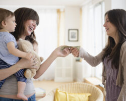 A mother holding a baby and a stuffed toy gives cash to a nanny in a bright, well-lit living room, symbolizing a pay raise for childcare services.
