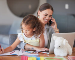 A family assistant multitasks by helping a young girl with homework while managing a phone call and working on a laptop, highlighting their role in supporting busy households.