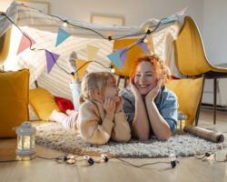 A smiling nanny and child lie on the floor inside a cozy blanket fort decorated with string lights and bunting, symbolizing the close bond and daily interaction often found in live-in nanny arrangements.