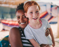 A joyful summer nanny embraces a laughing redheaded child on a sunny beachside deck, with hammocks swaying in the background, illustrating the warmth, fun, and personalized care a summer nanny provides