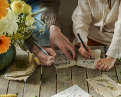 A child and adult decorate handmade pressed flower cards together at a wooden table, surrounded by flowers and craft supplies.