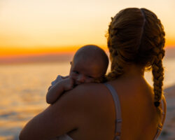 A baby nurse holds an infant at sunset during a family’s beach vacation, highlighting the benefits of traveling with a baby nurse.