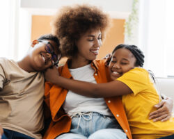 A nanny sits on a couch smiling as two children hug her tightly, symbolizing appreciation and love during National Nanny Recognition Week.