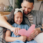 Father and daughter smiling on a couch while holding a handmade pink heart card, celebrating a kindness-focused Valentine’s Day activity together.