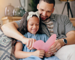 Father and daughter smiling on a couch while holding a handmade pink heart card, celebrating a kindness-focused Valentine’s Day activity together.