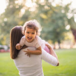 Summer caregiver lifting a joyful toddler in a park, symbolizing active and nurturing summer nanny support.