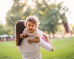 Summer caregiver lifting a joyful toddler in a park, symbolizing active and nurturing summer nanny support.