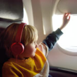 Child wearing headphones looks out airplane window, helping reduce sensory overload during travel