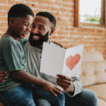 Father and child sharing a homemade Father’s Day card while spending quality time together at home, celebrating a simple and meaningful Father’s Day.