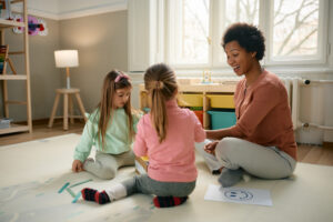 Happy governess and little girls having fun while sitting on the floor and playing.